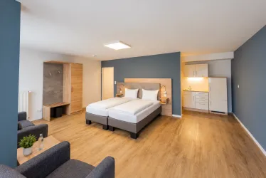 Ein modernes Zimmer mit einem Doppelbett und Holzfußboden. Die Ausstattung umfasst einen kleinen Wohnbereich und eine Küche.