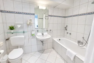 Ein helles Badezimmer mit weißen Fliesen und einem großen Spiegel. Es gibt eine Badewanne, ein Waschbecken und eine Toilette, sowie eine kleine Zimmerpflanze.