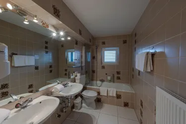 Ein modernes Badezimmer mit zwei Waschbecken, einer Badewanne und modernen Fliesen. Es gibt Fenster und beleuchtete Spiegel, die dem Raum eine helle Atmosphäre verleihen.