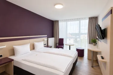 Ein modernes Hotelzimmer mit zwei Einzelbetten und einer großen Fensterfront. Der Raum ist in neutralen Tönen mit lila Akzenten gestaltet und bietet einen Fernseher sowie einen kleinen Tisch.