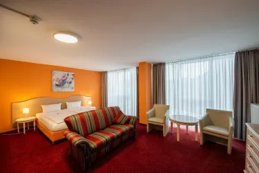 Ein modernes Hotelzimmer mit einem Doppelbett, einem bunten Sofa und zwei Stühlen. Große Fenster sorgen für viel Tageslicht und eine freundliche Atmosphäre.