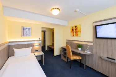Ein modernes Hotelzimmer mit einem Einzelbett, einem Schreibtisch und einem Fernseher. Die Wände sind in sanften Farben gestaltet, und es gibt einige Bilder zur Dekoration.