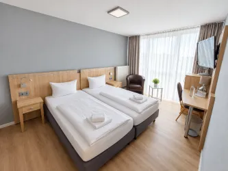 Ein modernes Hotelzimmer mit zwei Einzelbetten und großen Fenstern. Es gibt einen Schreibtisch und einen Sessel, der für eine angenehme Atmosphäre sorgt.