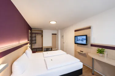 Ein modernes Hotelzimmer mit zwei Einzelbetten und einem Fernseher an der Wand. Die Einrichtung ist hell und freundlich gestaltet, mit einem Kleiderschrank und einem kleinen Tisch.