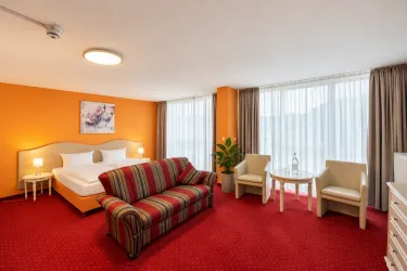 Ein gemütliches Zimmer mit rotem Teppich und orangefarbenen Wänden. Es gibt ein Doppelbett, einen bequemen Couchbereich und viel natürliches Licht durch große Fenster.