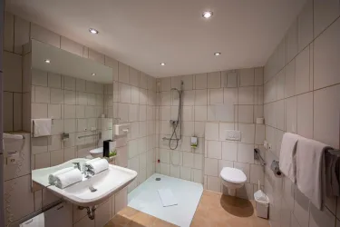 Ein modernes Badezimmer mit hellem Fliesenbelag und einer Dusche. Es gibt ein Waschbecken, eine Toilette und verschiedene Handtücher.