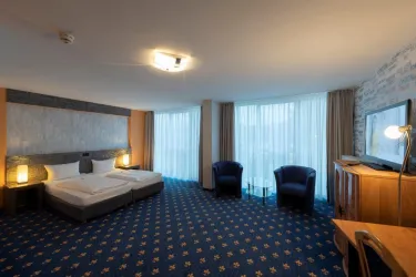 Ein modernes Hotelzimmer mit zwei Betten, blauen Sessel und einem Tisch. Große Fenster lassen viel Licht hinein und bieten einen Blick nach außen.