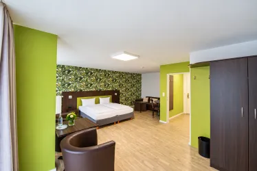 Ein modernes Hotelzimmer mit grünen Wänden und einer musterbedeckten Wand. Es gibt ein Bett, einen Tisch und einen Stuhl sowie einen Kleiderschrank.