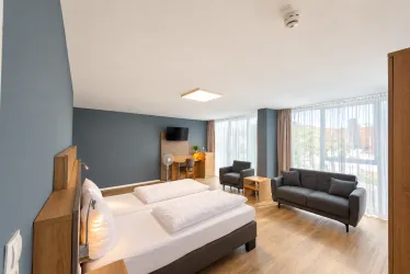 Ein modernes Schlafzimmer mit einem großen Bett und einem Sofa. Die Fenster lassen viel Tageslicht herein und bieten einen Blick nach draußen.