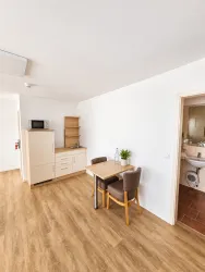 Ein helles Wohn- und Esszimmer mit einem Tisch und zwei Stühlen. Auf der linken Seite befindet sich eine Kücheneinheit mit einem Mikrowellenherd und offenen Regalen.