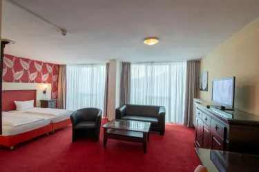 Ein modernes Hotelzimmer mit einem Doppelbett und einem gemütlichen Sitzbereich. Große Fenster lassen viel Tageslicht herein und sorgen für eine helle Atmosphäre.