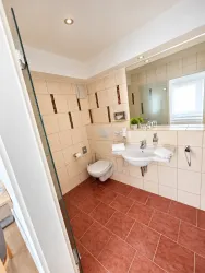 Ein modernes Badezimmer mit hellen Fliesen und einer großen Duschkabine. Es enthält ein Waschbecken, eine Toilette und natürliche Lichtquellen.