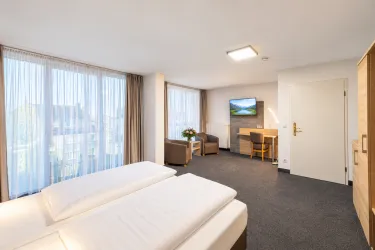 Ein modernes Hotelzimmer mit zwei Einzelbetten und großen Fenstern. Der Raum ist hell und freundlich eingerichtet, mit einem Tisch und einem Fernseher.
