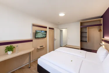 Ein modernes Hotelzimmer mit zwei Einzelbetten und einem Flachbildfernseher. Der Raum ist hell und schlicht eingerichtet, mit einem minimalistischen Schreibtisch und Stauraum.