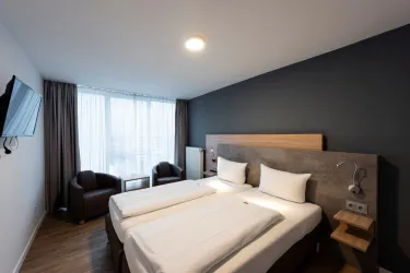 Ein modernes Hotelzimmer mit zwei Einzelbetten und einem Fernseher. Der Raum hat große Fenster, gemütliche Stühle und eine schlichte, elegante Gestaltung.