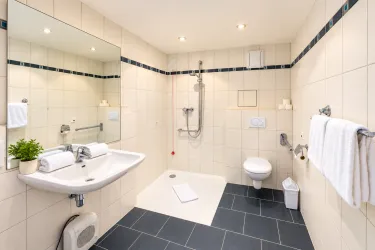 Ein modernes Badezimmer mit einer Dusche, einem Waschbecken und einem WC. Die Wände sind hell gefliest, und es gibt handtücher und Pflanzen zur Dekoration.