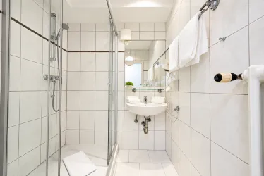 Ein modernes Badezimmer mit einer Duschkabine und einem Waschbecken. Weiße Fliesen und Handtücher sorgen für ein sauberes Ambiente.