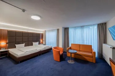 Ein modernes Hotelzimmer mit zwei Einzelbetten und einem gemütlichen Wohnbereich. Große Fenster lassen viel Tageslicht herein und der Teppichboden ist dunkelblau.