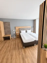 Ein modernes Hotelzimmer mit zwei Einzelbetten und einem großen Fenster. Der Boden ist aus Holz, und es gibt einen Ventilator sowie eine Holzbank.