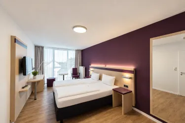 Ein modernes Hotelzimmer mit einem Doppelbett und einem großen Fenster. Die Wände sind in einem eleganten Violettton gestrichen, und die Einrichtung ist stilvoll und minimalistisch.