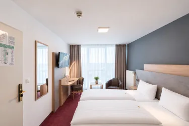 Ein modernes Hotelzimmer mit zwei Einzelbetten und einem Schreibtisch. Große Fenster sorgen für viel Licht und eine angenehme Atmosphäre.