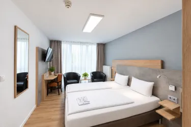 Ein modernes Hotelzimmer mit zwei Einzelbetten und einem großen Fenster. Es gibt einen Schreibtisch, Stühle und einen Fernseher.