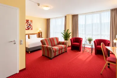 Ein helles Hotelzimmer mit einem roten Teppich und großen Fenstern. Es gibt ein Bett, einen gemütlichen Sofa und zwei Sessel.
