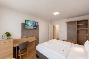 Ein modernes Hotelzimmer mit einem Doppelbett, einem Schreibtisch und einem Fernseher. Die Wände sind hell und die Einrichtung ist schlicht und elegant.