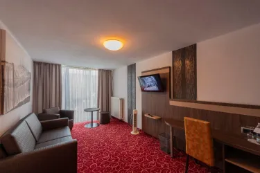 Ein modernes Hotelzimmer mit einem komfortablen Sofa, einem Tisch und einem Fernseher. Große Fenster sorgen für viel Tageslicht.