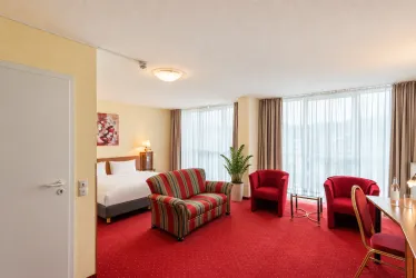 Ein gemütliches Hotelzimmer mit rotem Teppich und modernen Möbeln. Die großen Fenster bieten viel Tageslicht und einen Blick nach draußen.