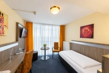 Ein modernes Hotelzimmer mit einem Einzelbett und einem Tisch. Große Fenster lassen viel Tageslicht herein und es gibt dekorative Kunst an den Wänden.