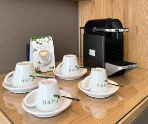 Eine Kaffeemaschine steht neben vier Tassen auf einem Holztisch. Dazu liegt eine Broschüre für die verwendeten Kaffeekapseln.