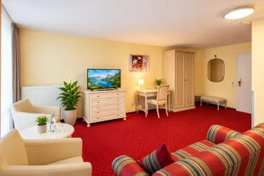 Ein helles und gemütliches Zimmer mit rotem Teppich, einer Couch und einem Fernseher. Es gibt einen Schreibtisch und eine Pflanze, die dem Raum Leben verleiht.