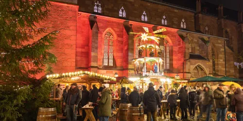 Eine festliche Szene mit einem Weihnachtsmarkt bei Nacht. Menschen stehen in der Nähe von Ständen mit Lichtern und Dekorationen.
