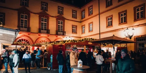 Ein festlicher Weihnachtsmarkt mit beleuchteten Ständen und Menschen, die sich umsehen. Die Atmosphäre ist gemütlich und einladend, umgeben von historischen Gebäuden.