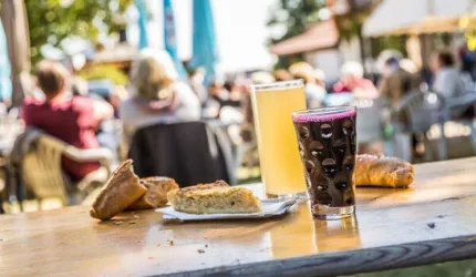 Ein Tisch mit verschiedenen Snacks und Getränken, darunter ein Glas Bier und ein Getränk in einem hohen Glas. Im Hintergrund sind Menschen zu sehen, die die entspannte Atmosphäre genießen.