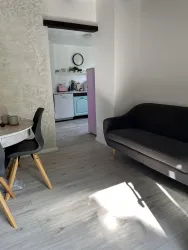 Ein heller Wohnraum mit einem grauen Sofa und einem kleinen Esstisch. Im Hintergrund ist eine Küche mit bunten Möbeln sichtbar.