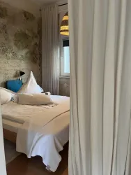 Ein gemütliches Schlafzimmer mit einem großen Bett und hellem Bettzeug. An der Wand sind rustikale Steinstrukturen und Vorhänge, die den Raum von einem Flur abtrennen.