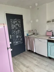 Eine moderne Küche mit einem rosa Kühlschrank und einem hellblauen Geschirrspüler. Die Wände sind hell und es gibt eine schöne schwarze Tür mit einer dekorativen Zeichnung.