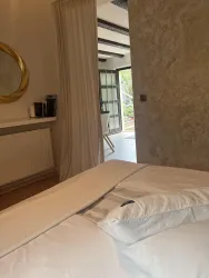 Ein gemütliches Schlafzimmer mit einem weißen Bett und einem großen Spiegel. Durch die Fenster sieht man einen lichtdurchfluteten Raum mit Zugang nach draußen.