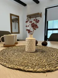 Ein stilvoller Tisch mit einer geflochtenen Tischdecke, einer kleinen weißen Vase und getrockneten Blumen. Im Hintergrund sind eine Tasse und ein Spiegel zu sehen.