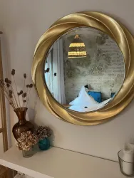 Ein eleganter Spiegel mit goldenen Details hängt an der Wand. Im Spiegel ist eine entspannende Umgebung mit einem Bett und einer Lampe zu sehen.