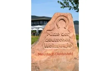 Ein Wegweiser aus Stein mit der Aufschrift "Mitte der deutschen Weinstraße". Im Hintergrund sind Bäume und ein Gebäude sichtbar.