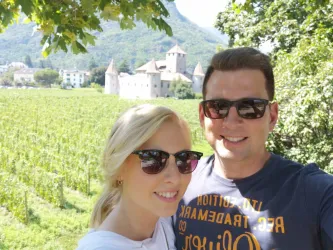 Ein Paar posiert lächelnd vor einer malerischen Landschaft mit Weinbergen und einer Burg im Hintergrund. Der Himmel ist klar und die Natur ist üppig grün.