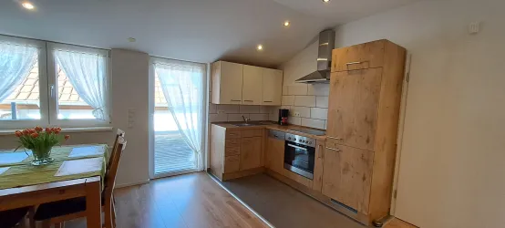 Eine moderne Küche mit Holzschränken und einem Herd. Große Fenster lassen viel Licht herein und bieten Zugang zum Außenbereich.