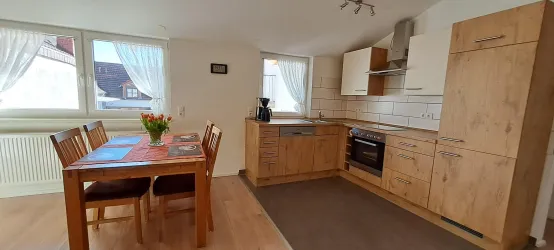 Eine moderne Küche mit einer Essgruppe aus Holz und großen Fenstern. Der Raum ist hell und freundlich mit einer gemütlichen Atmosphäre.