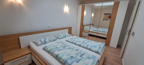 Ein modernes Schlafzimmer mit einem Doppelbett und hellen Wänden. Der Raum verfügt über einen großen Spiegel und Holzelemente, die eine gemütliche Atmosphäre schaffen.