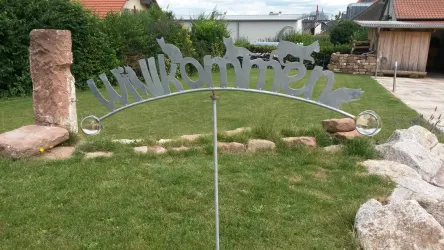 Ein Willkommensschild aus Metall steht in einem Garten. Der Hintergrund zeigt eine grüne Wiese und eine einfache, ländliche Umgebung.