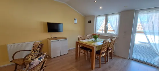 Ein gemütlicher Wohn- und Essbereich mit gelben Wänden und einem Esstisch für mehrere Personen. Große Fenster sorgen für viel Tageslicht und einen Blick nach draußen.