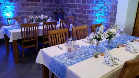 Unsere Weinstube festlich dekoriert. Ein eleganter Raum mit Holzstühlen und weißen Tischen, dekoriert mit blauen Tischläufern. Kleine Blumenarrangements und Gläser ergänzen die ansprechende Atmosphäre.